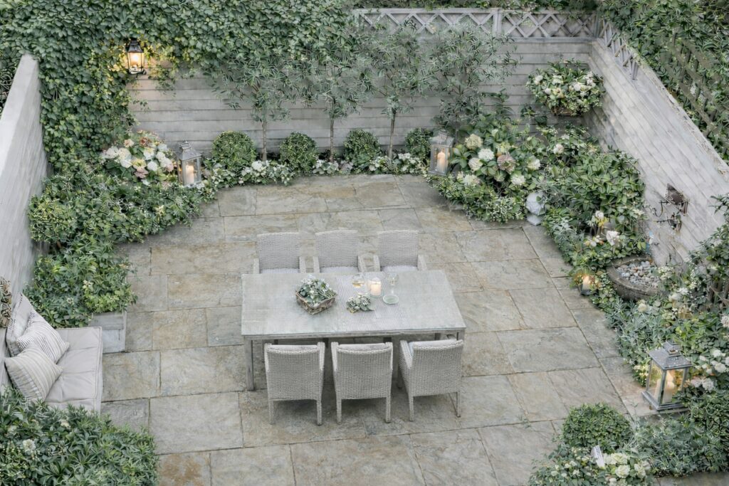 A small, landscaped patio garden, enclosed, including a dining table set surrounded by lush vegetation, including shrubs, potted plants and small trees. This landscaped space has a light stone floor and is surrounded by wooden fences. The landscaping company has designed this landscaping service to create a soothing and harmonious atmosphere.